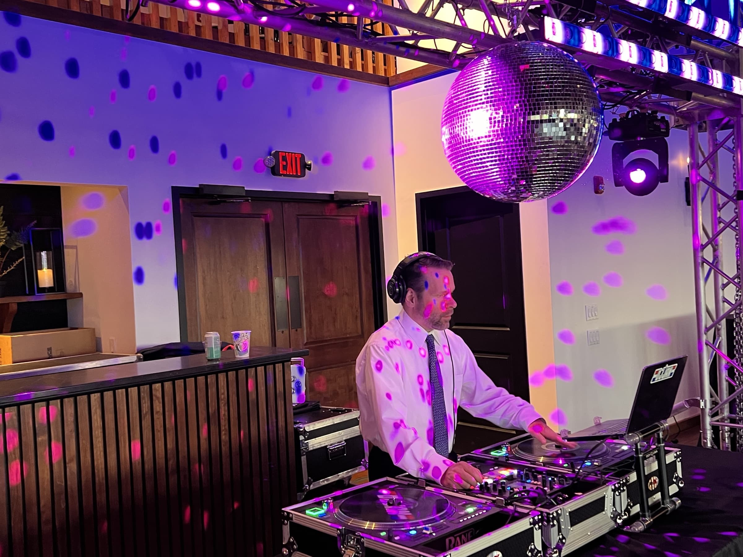 DJ in headphones on the turntables beneath a mirror ball and truss-mounted moving head, the room dotted purple