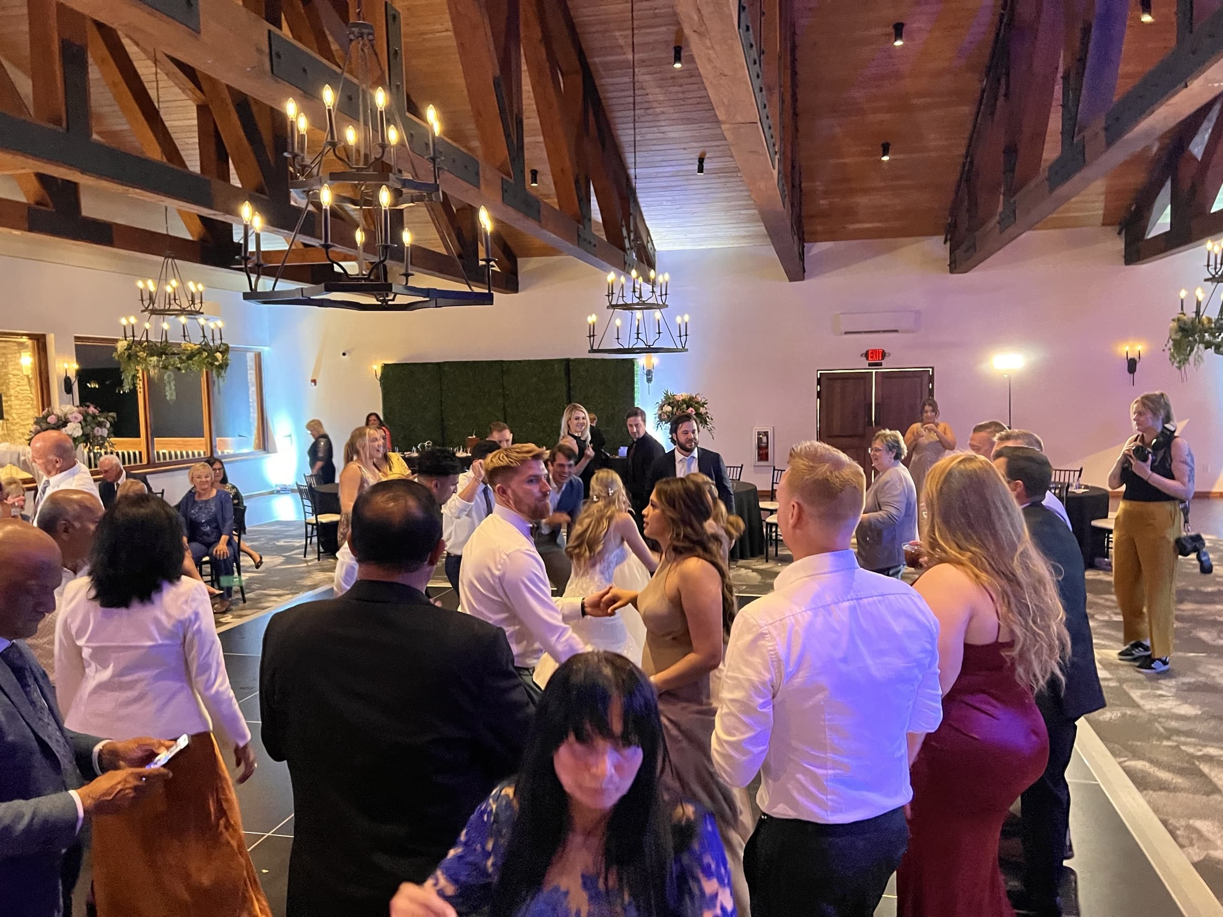 Wide view of a wedding reception dance floor beneath vaulted timber beams and iron chandeliers