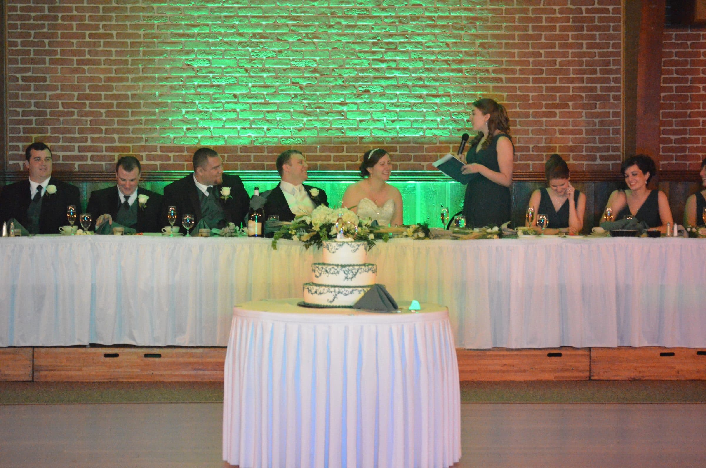 Maid of honor gives a toast at the head table, a brick wall behind the wedding party washed in green uplighting