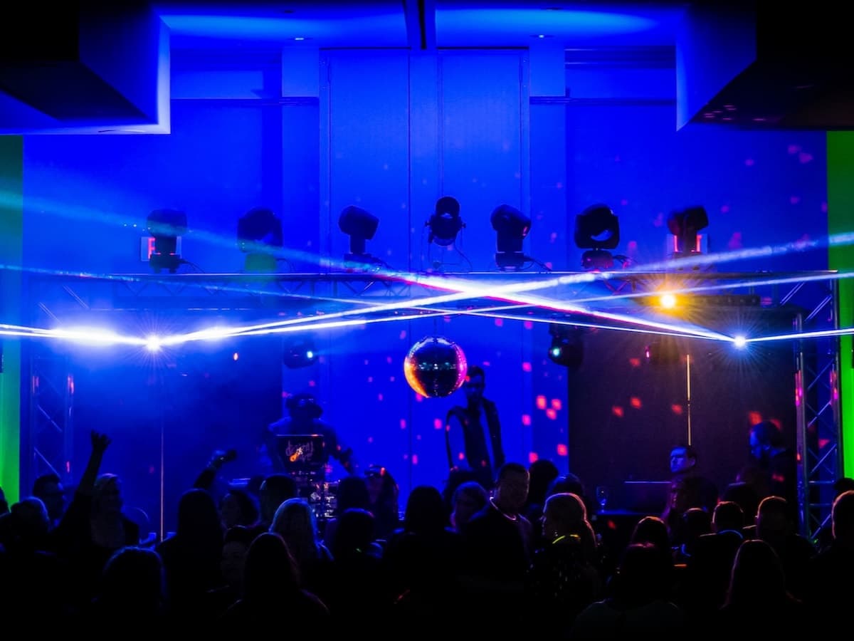 Two laser beams cross above a blue-lit dance floor under a truss rig and mirror ball