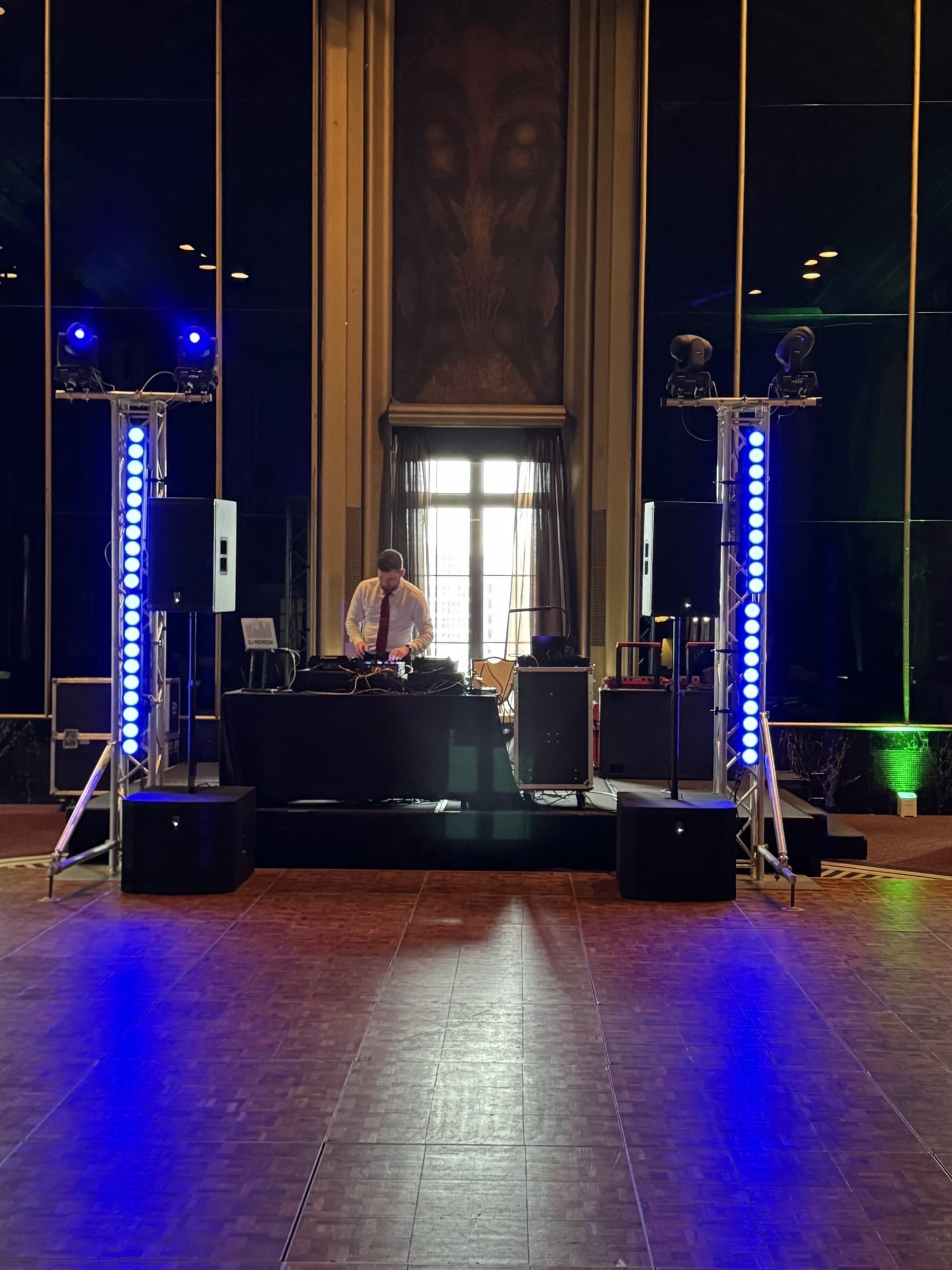 Gold setup: DJ between two lighting totems, each with moving heads and a vertical LED bar, in a tall ornate hall