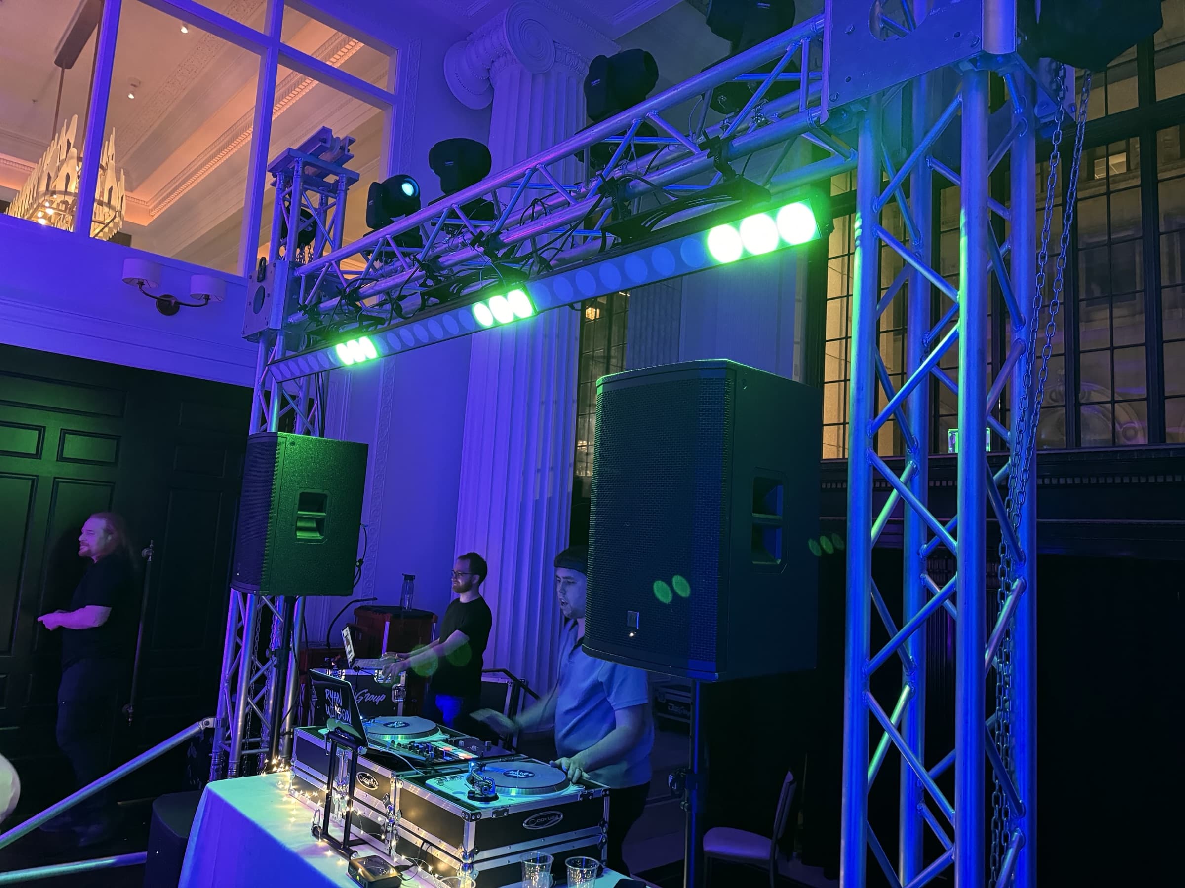 Gold setup: goalpost truss with moving heads and LED bars lit blue over the DJ in a columned downtown venue
