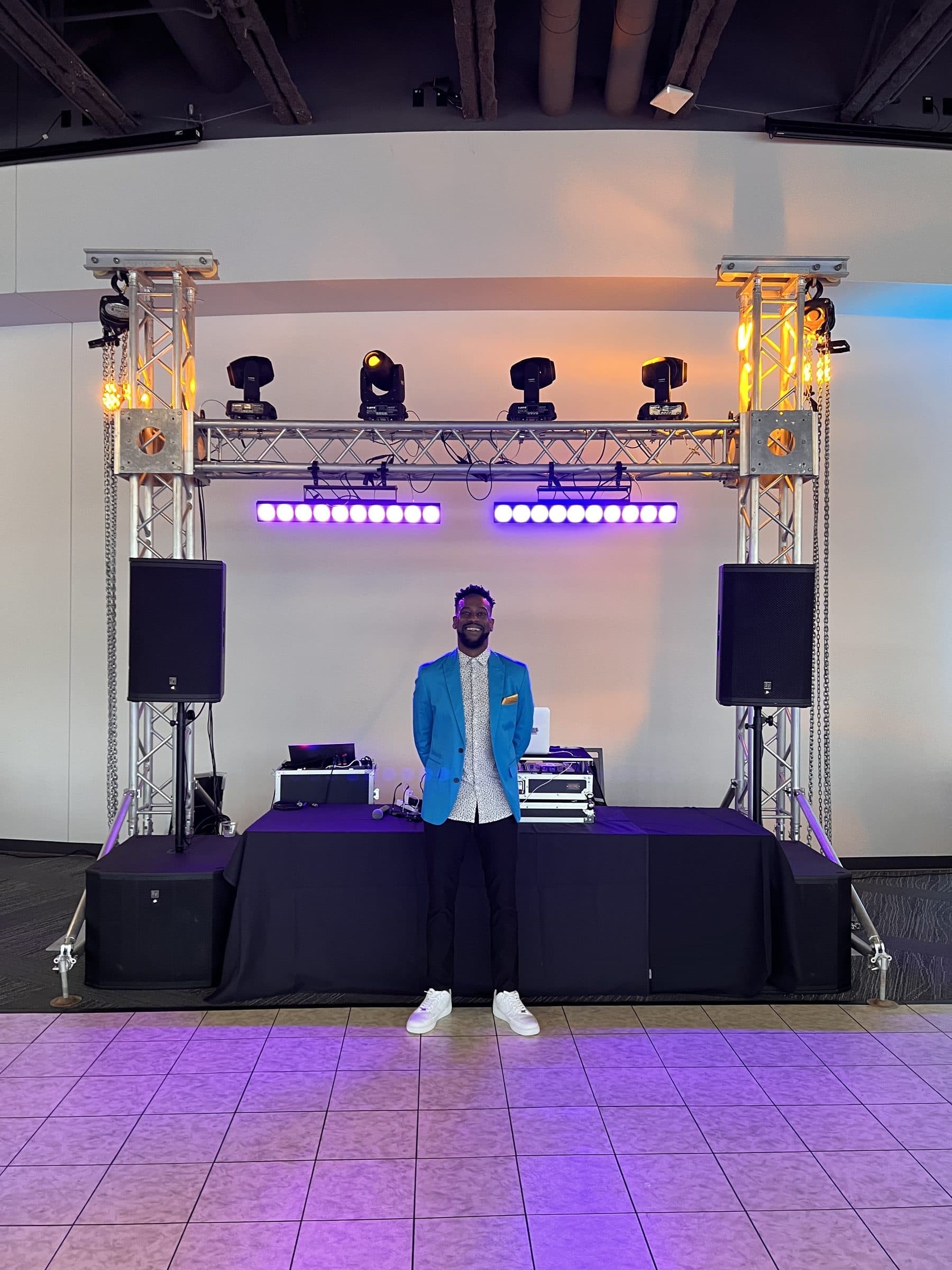 Gold setup: DJ in a blue blazer in front of a goalpost truss with four moving heads, twin LED bars and amber uplit towers
