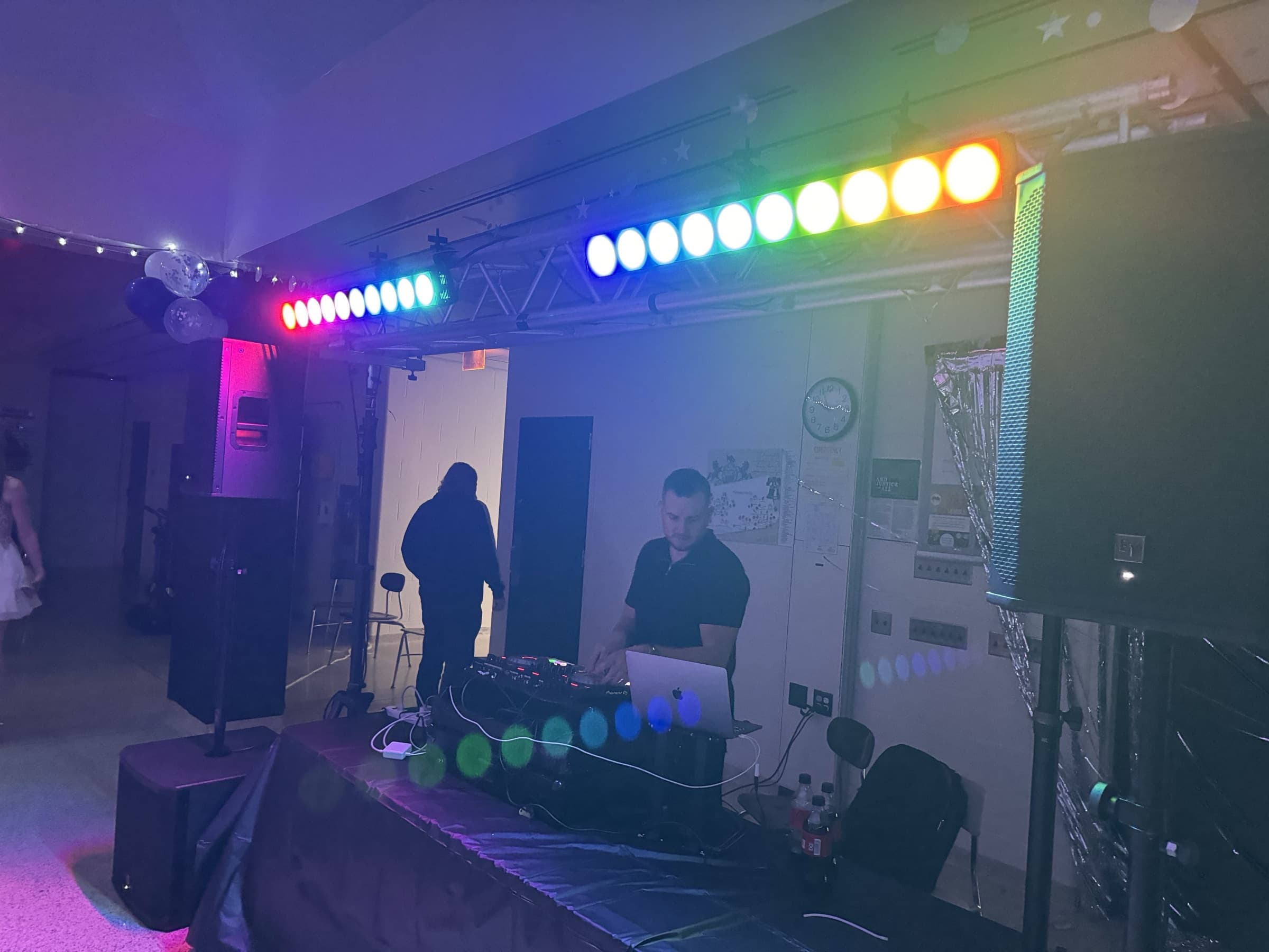 Silver setup at a school dance: rainbow LED bars on a crossbar above the DJ, speakers on stands either side