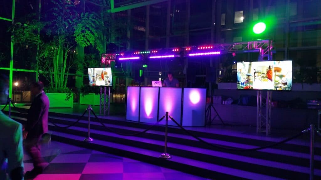 DJ setup with truss-mounted LED bars and twin screens in a glass atrium at a corporate event
