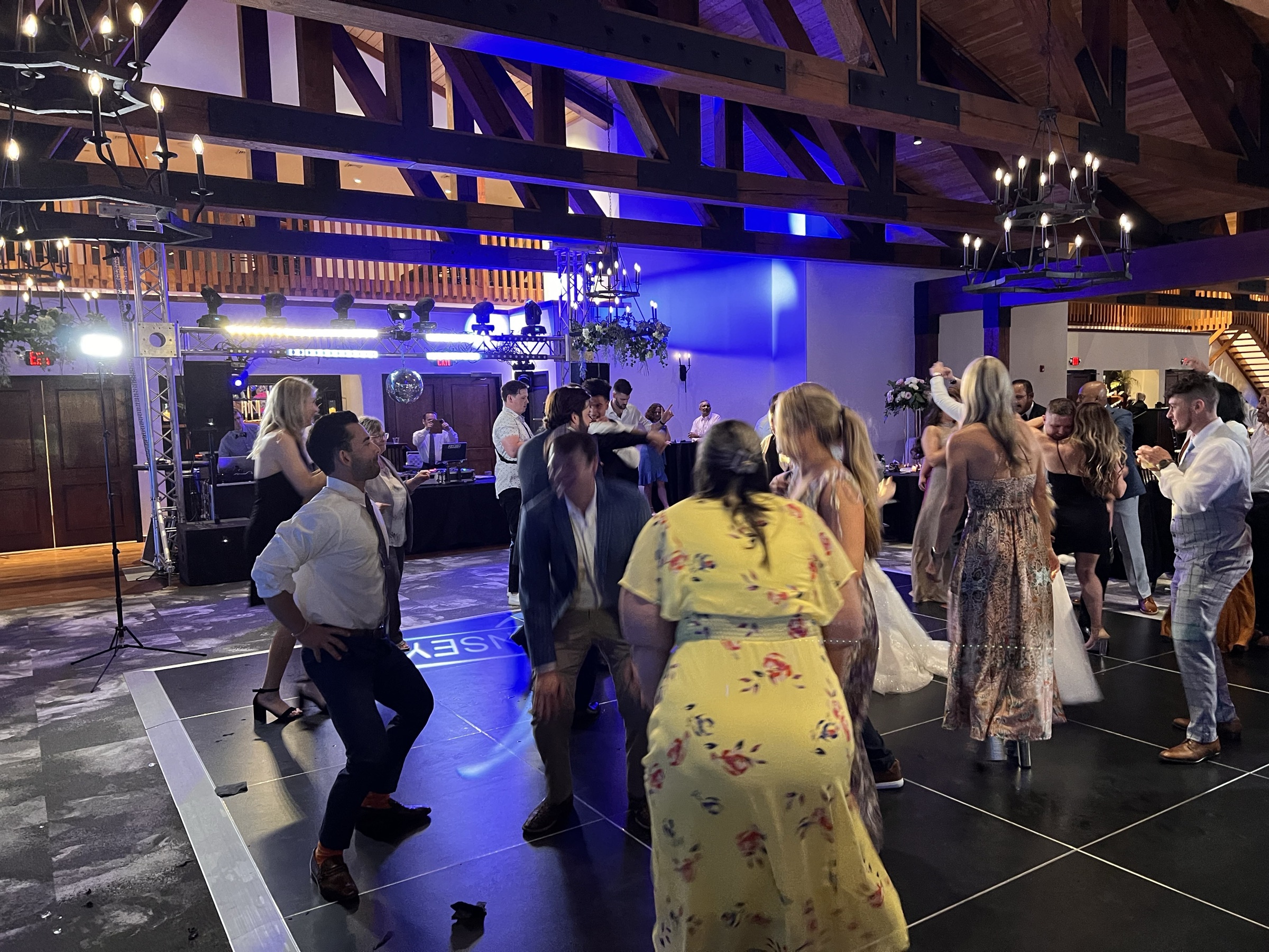 Wedding guests dance over the couple's monogram projected on a black dance floor, the bride's gown at right, truss lighting and blue uplighting under timber beams