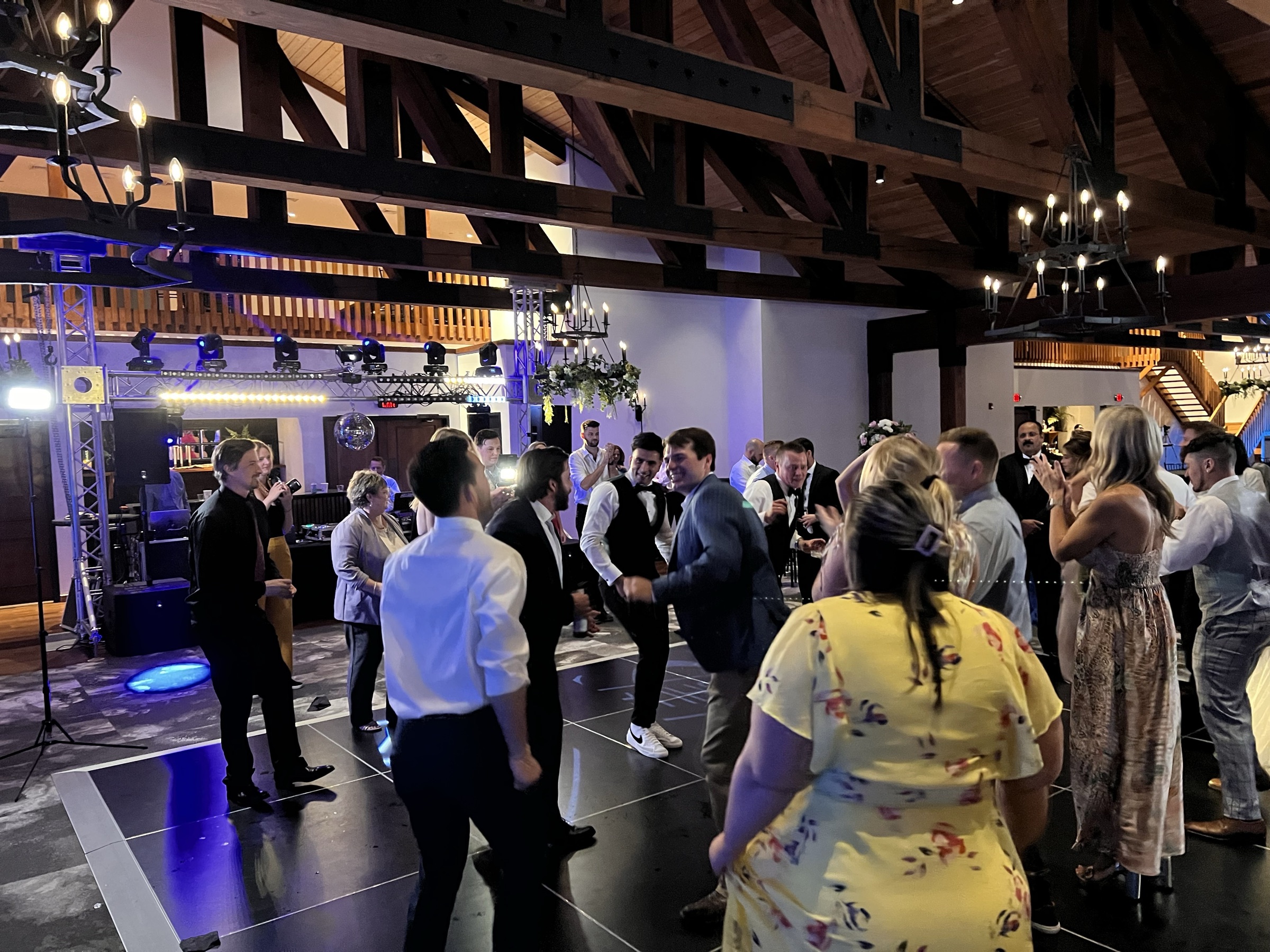 Groomsmen in vests dance in a ring of guests beneath the truss rig and mirror ball at a wedding reception