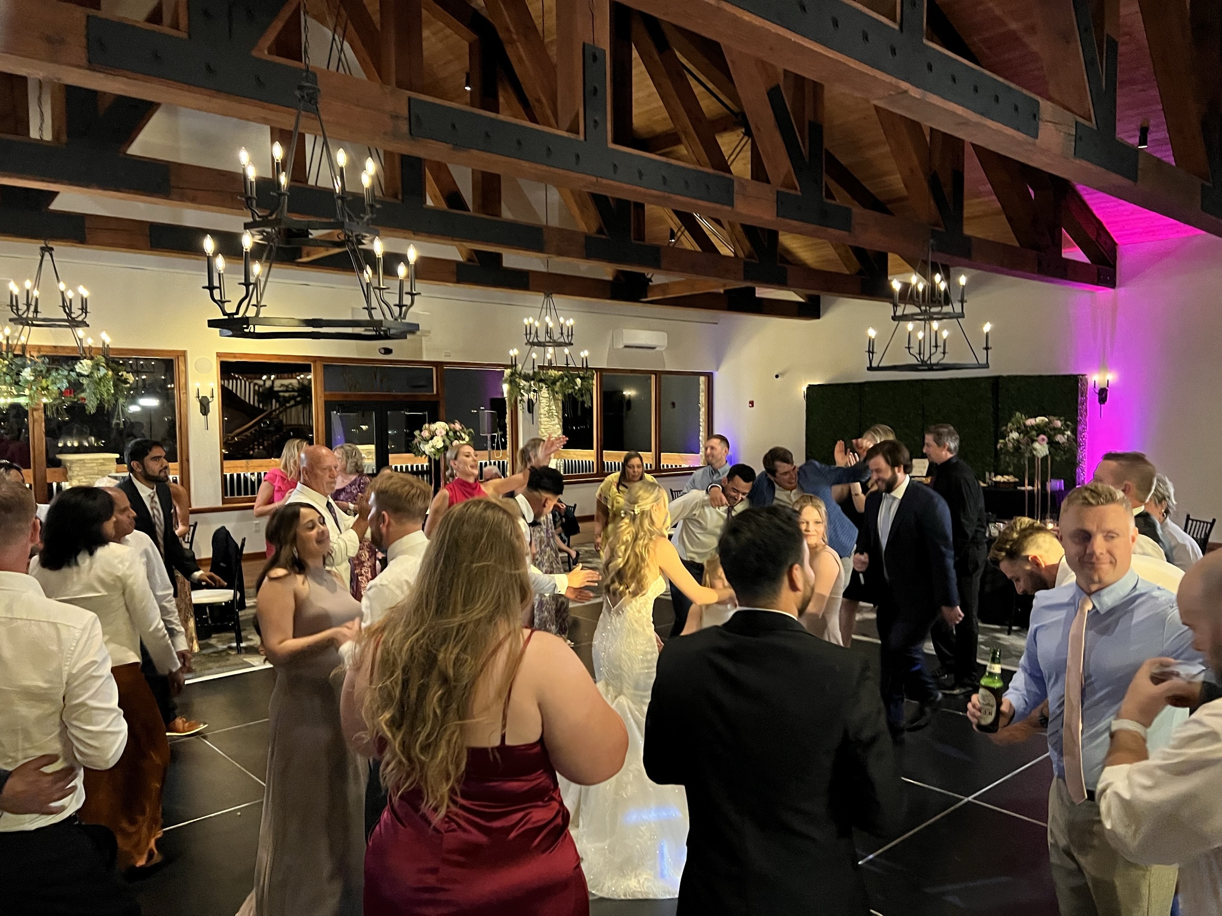 Bride dances at the center of a circle of guests beneath iron chandeliers, magenta uplighting on the wall behind
