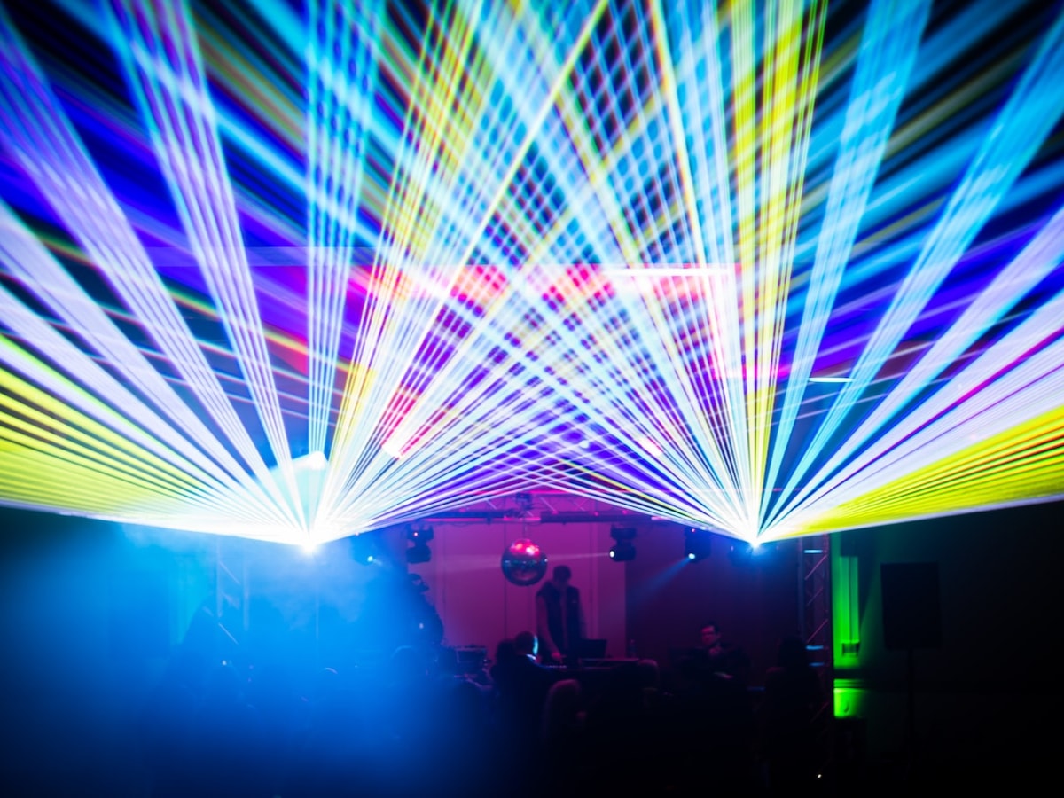 Blue and yellow laser beams fan over the truss rig, mirror ball and DJ in a hazy ballroom