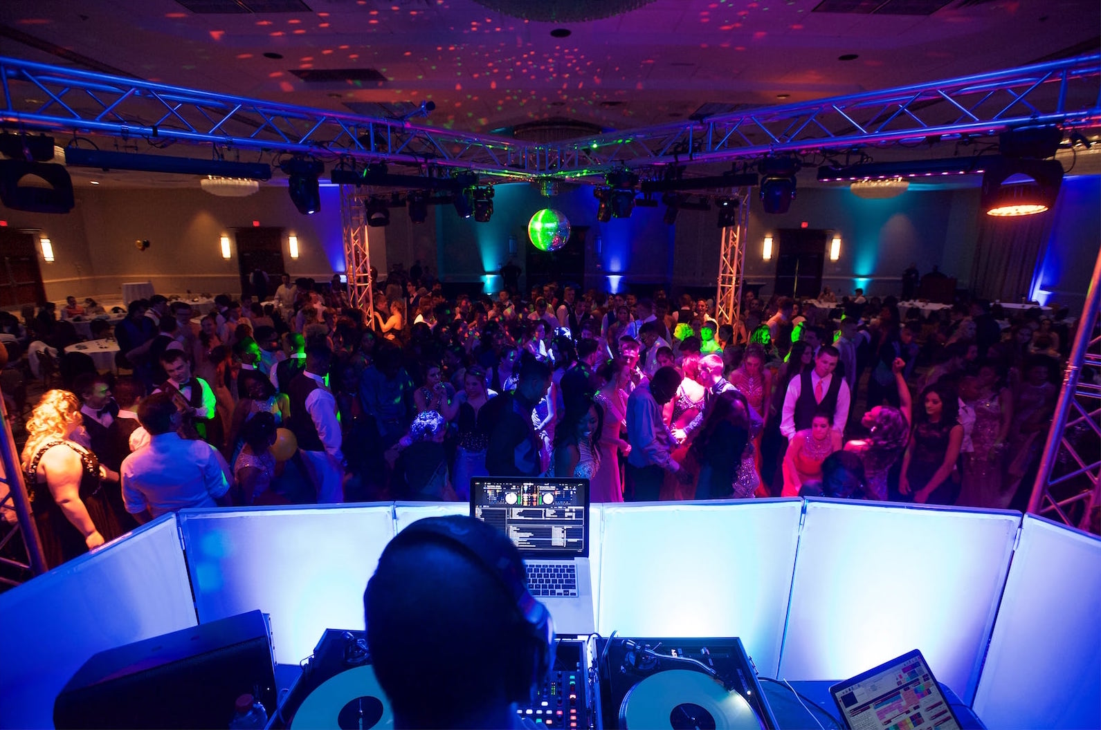 Packed prom dance floor seen from the DJ booth under an X-shaped truss rig and mirror ball