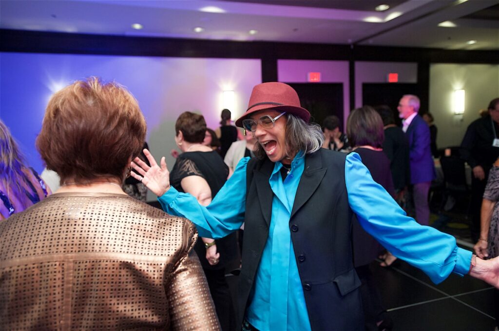 A guest in a red fedora dances with arms wide at a celebration in a Pittsburgh ballroom